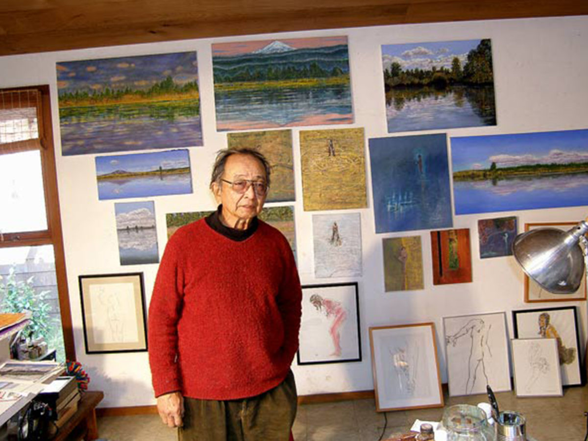 Old man in a red sweater stands in front of a wall hung with many sketches and landscape paintings.